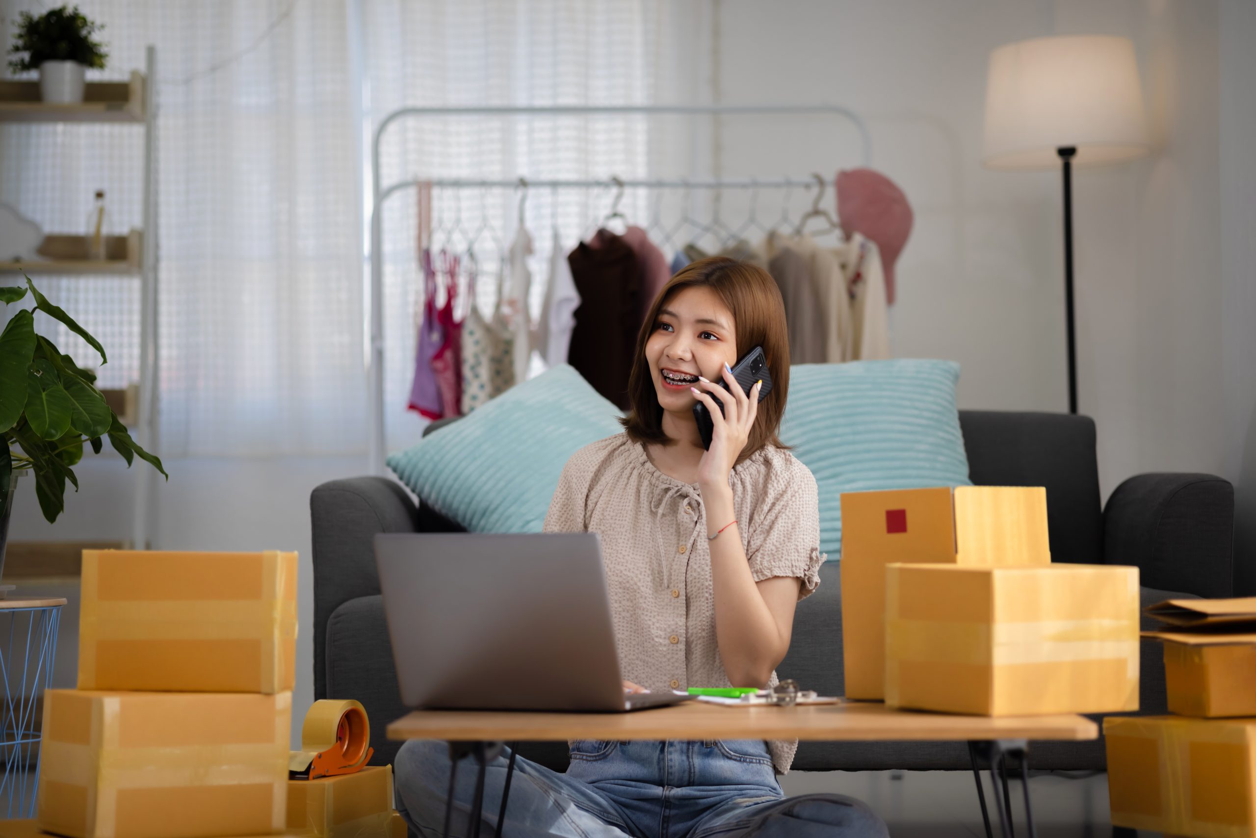 smiling happy young asian woman entrepreneur receiving phone call new sales order among boxes product with laptop computer scaled